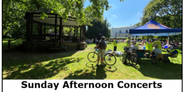 Crowds relax on grass in a sunny park while listening to a live band on a bandstand, with event tents and trees in the background.