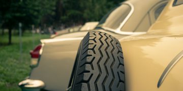 Close-up of a classic car’s spare tire mounted on the side, with another vintage car in the background.