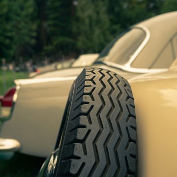 Close-up of a classic car’s spare tire mounted on the side, with another vintage car in the background.