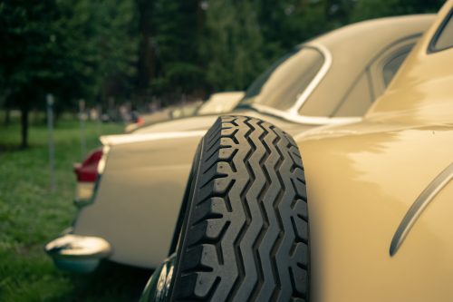 Close-up of a classic car’s spare tire mounted on the side, with another vintage car in the background.