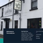White building with "The Lamb Inn" pub sign, a silver car parked in front, and opening times displayed on a blue banner hanging on the wall.