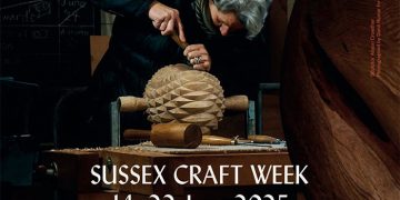 Older woman carving a wooden sculpture at a table with Sussex Craft Week, 14-22 June 2025, West Dean, advertised in the background.