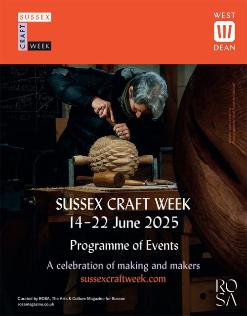 Older woman carving a wooden sculpture at a table with Sussex Craft Week, 14-22 June 2025, West Dean, advertised in the background.