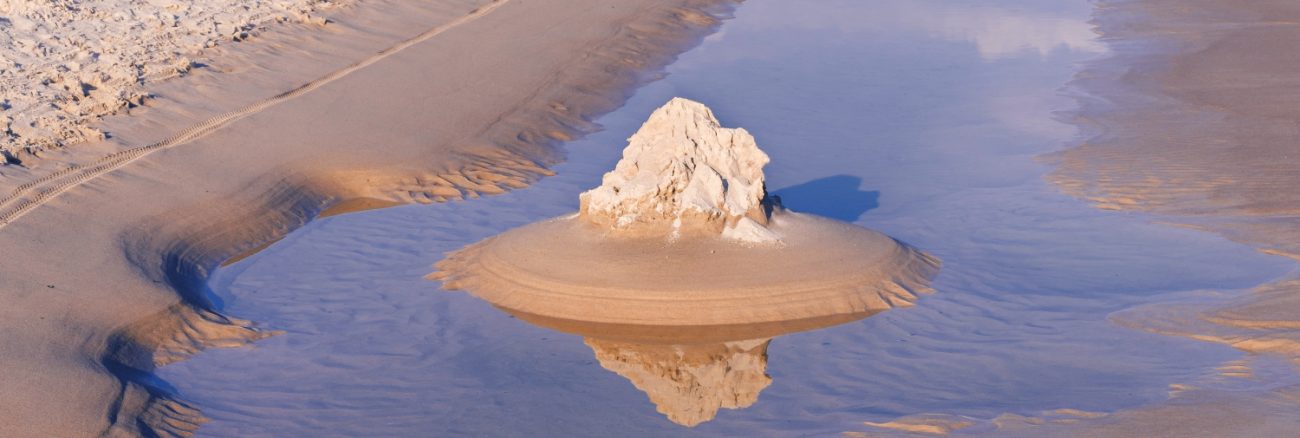 A small sand formation is reflected in shallow water on a beach.